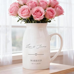 Op de afbeelding: Een witte keramische vaas met een handvat, gevuld met roze rozen. De vaas heeft de namen "Lisa & Travis" en het woord "GETROUWD" met een datum eronder. De vaas staat op een houten oppervlak bij een raam.