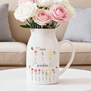 Puede incluir: Una jarra de cerámica blanca llena con un ramo de rosas blancas y rosas. El frente de la jarra presenta el texto "FIRST mom" sobre tres ilustraciones de flores con nombres, y "NOW grandma" sobre seis ilustraciones de flores con nombres.