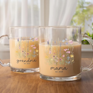 May include: Two clear glass mugs with handles, each filled with a light brown liquid. One mug says "grandma est. 2020" and the other says "mama est. 2025". Both mugs feature a floral design with pink, purple, and yellow flowers.