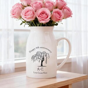 Könnte beinhalten: Eine weiße Keramikvase mit Henkel, gefüllt mit rosa Rosen. Die Vase hat eine schwarze Grafik einer Trauerweide und den Text "Happy 9th anniversary", "A+E" und "Love Grows Here". Die Vase steht auf einem Holztisch.