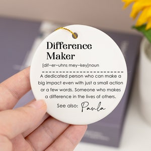 Pode incluir: Ornamento de cerâmica branca com as palavras "Difference Maker" e uma definição da palavra. A definição diz: "Uma pessoa dedicada que pode ter um grande impacto mesmo com uma pequena ação ou poucas palavras. Alguém que faz a diferença na vida dos outros." O ornamento também inclui o texto "Veja também: Paula".