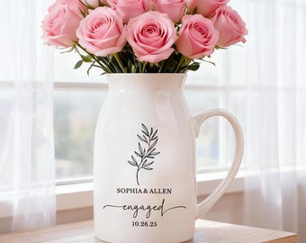 May include: A white ceramic pitcher vase filled with a bouquet of light pink roses. The front of the pitcher features a black line drawing of a leafy branch above the text "SOPHIA & ALLEN", "engaged", and the date "10.26.25".