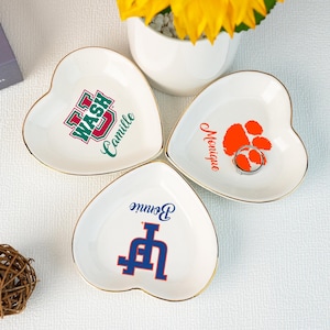 May include: Three white heart-shaped ceramic trinket dishes with gold trim. Each dish has a different college logo and a name. The first dish has the University of Washington logo and the name "Camille". The second dish has the Clemson University logo and the name "Monique". The third dish has the University of Louisville logo and the name "Bonnie".