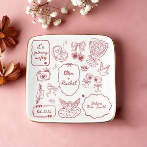 May include: Square white ceramic plate with a gold rim, decorated with red line art illustrations of wedding symbols and text. Includes a ring, flowers, and the names "Ellen + Rachel" and "Future Mrs. Smith."
