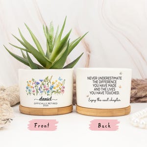 Puede incluir: Dos macetas de cerámica blanca con platillos de bambú, cada una con una planta suculenta. Una maceta presenta un diseño floral y el nombre "daniel" con "OFFICIALLY RETIRED 2025". La otra dice "NEVER UNDERESTIMATE...Enjoy the next chapter."