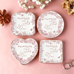 May include: Four white ceramic plates with gold rims, each featuring a floral and butterfly design in a reddish-brown hue. The plates are square, round, heart-shaped, and rectangular, with the text "Always under the same sky" and names "Luna" and "James".