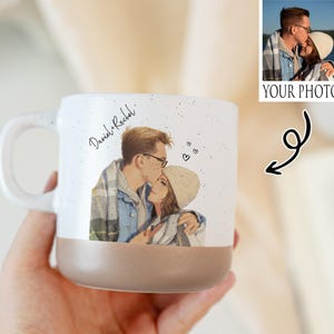 Puede incluir: Una taza de cerámica blanca con un acabado moteado y una base marrón. La taza presenta una ilustración personalizada de una pareja besándose, con los nombres "Daniel & Rachel" escritos encima. Se incluye una foto pequeña.