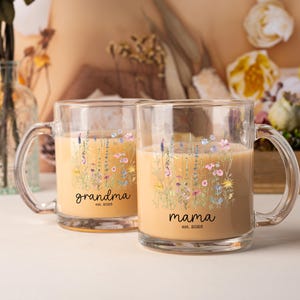 May include: Two clear glass mugs with handles, filled with a light brown liquid. Each mug features a floral design with the words "grandma est. 2025" and "mama est. 2025" in black script. The mugs are decorated with colorful wildflowers.