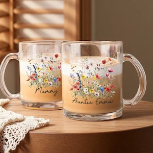 Puede incluir: Dos tazas de vidrio transparente con asas, cada una llena de una bebida cremosa. Las tazas están decoradas con un diseño floral y las palabras "Mummy" y "Auntie Emma". Las tazas están sobre una mesa de madera.