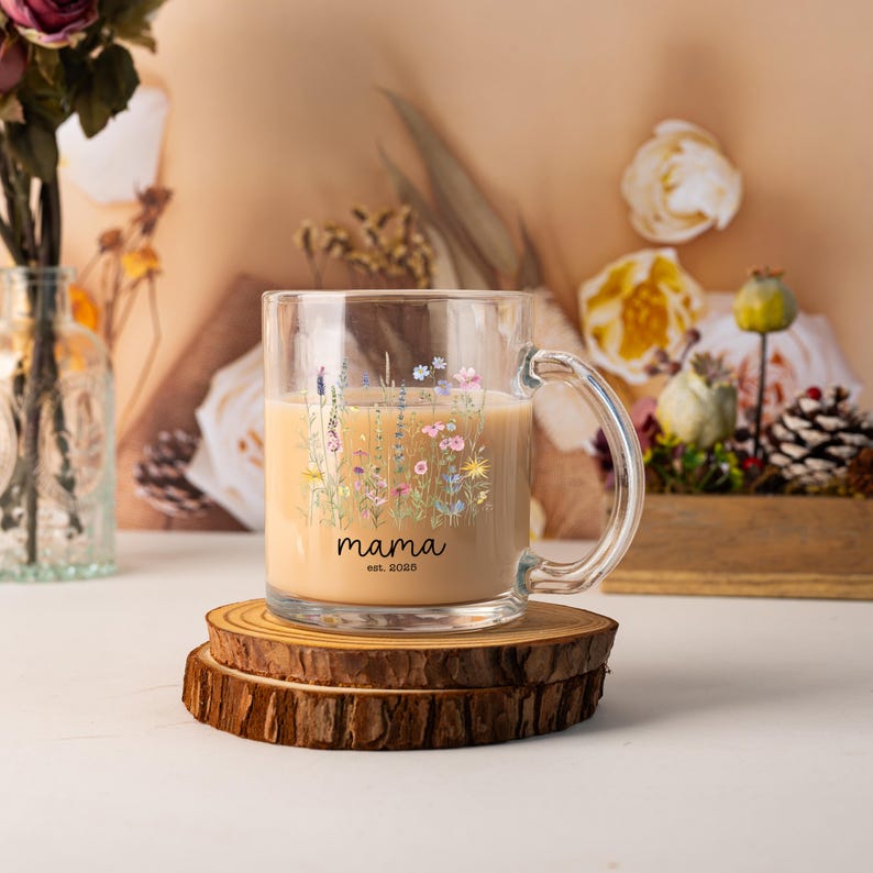 May include: A clear glass mug filled with a light-colored beverage, featuring a floral design and the word "mama" with "est. 2025" printed below. The mug sits on a wooden coaster, with a blurred background of flowers.