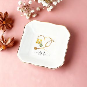 Op de afbeelding: Witte vierkante keramische schaal met een gouden rand en een gouden stethoscoopontwerp met gele bloemen en de naam "Chloe" in zwart schrift. De schaal heeft een golvende rand op een roze achtergrond.
