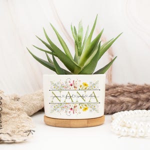 May include: White ceramic planter with a floral design and the word "NANA" in gold lettering. The planter has a wooden base and is filled with a green succulent plant. The planter also has the names "Joanna,  Furqan, Randy, Oliver" written in gold lettering.