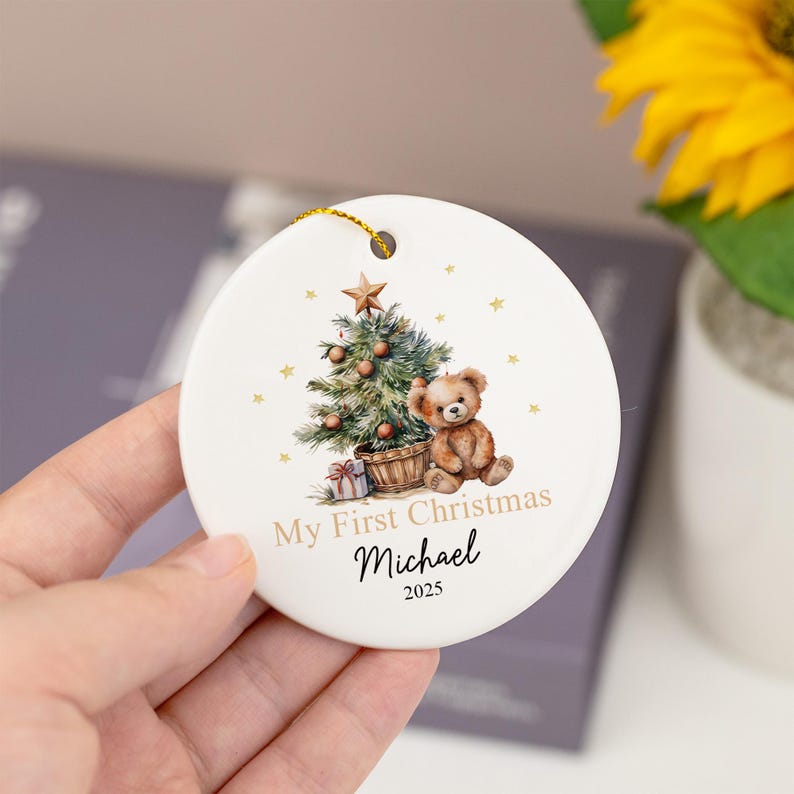 May include: A round white ceramic ornament featuring a printed illustration of a teddy bear sitting next to a decorated Christmas tree with a star on top. The text reads My First Christmas, the name Michael, and the year 2025 in black font.