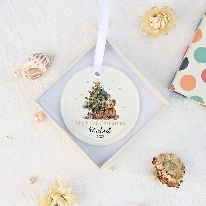 May include: A round white ceramic ornament hanging by a white ribbon, featuring a printed illustration of a decorated Christmas tree, a teddy bear, and gift boxes. The text reads My First Christmas, Michael, and the year 2025 in black font.