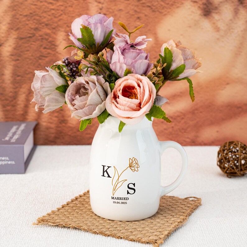 May include: White ceramic vase with a gold floral design and the initials "K" and "S" with the text "Married 19.04.2023". The vase is filled with a bouquet of pink and purple artificial flowers.