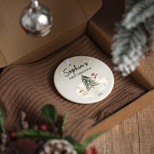 May include: A round, off-white Christmas ornament with the text "Sophia's First Christmas" and a winter scene with a bunny, snowman, and Christmas tree. The ornament is in a cardboard box on a brown knitted surface.