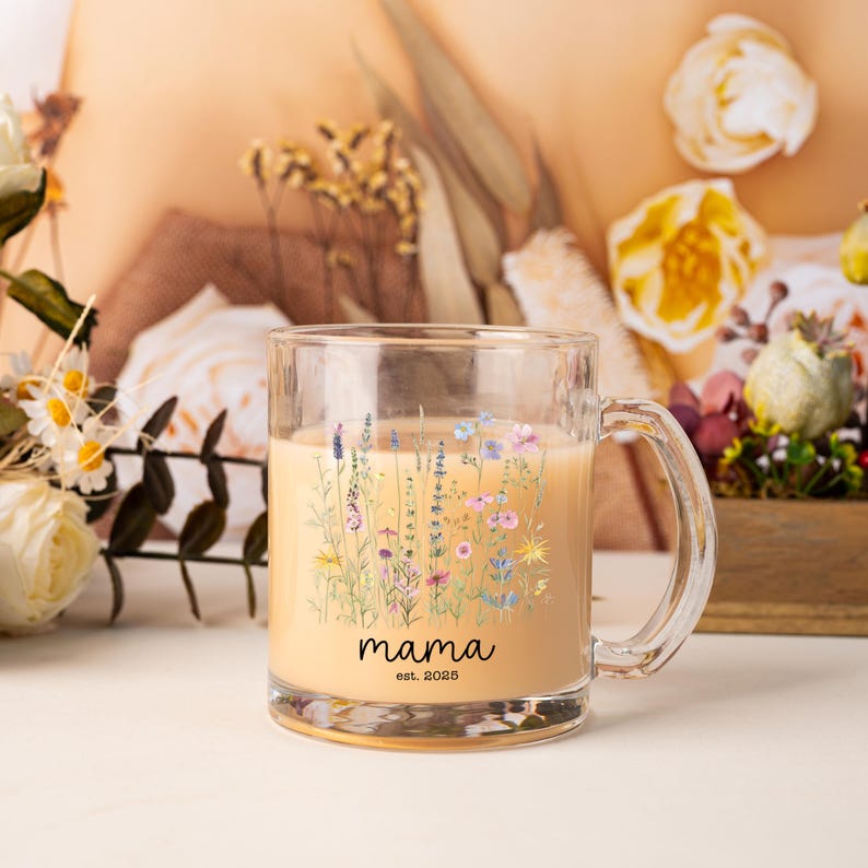 May include: A clear glass mug filled with a light-colored beverage. The mug features a floral design with the word "mama" and the year "est. 2023" printed in black. The background includes flowers and a wooden box.