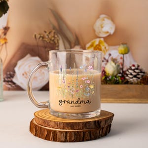 May include: A clear glass mug filled with a light brown liquid, decorated with a floral design in pastel colors. The mug has the word "grandma" and "est. 2025" printed on it. The mug sits on a wooden coaster.