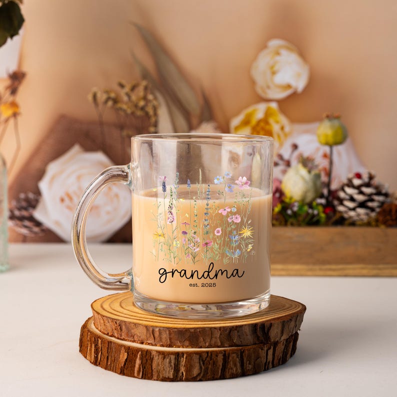 May include: A clear glass mug filled with a light brown liquid, decorated with a floral design in pastel colors. The mug has the word "grandma" and "est. 2025" printed on it. The mug sits on a wooden coaster.