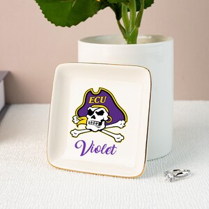 Puede incluir: Un plato cuadrado de cer&aacute;mica blanquecina con borde dorado. El plato presenta un logotipo pirata morado y dorado con el texto "ECU" y el nombre "Violet" en escritura morada. Un anillo est&aacute; cerca.