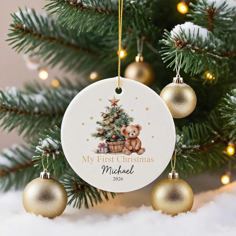 May include: A round white ceramic ornament featuring a watercolor illustration of a Christmas tree and a teddy bear. The text reads My First Christmas, Michael, and 2026. It hangs from a gold string against a background of green pine branches.