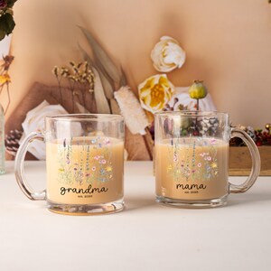 May include: Two clear glass mugs with floral designs and the words "grandma" and "mama" printed on them. The mugs are filled with a light-colored liquid and have a clear handle. The text "est. 2022" is printed below the words.