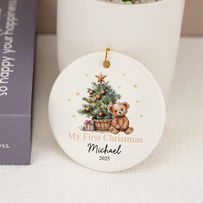 May include: A round white ceramic ornament featuring a watercolor illustration of a decorated Christmas tree, a brown teddy bear, and a gift box. The text reads My First Christmas, the name Michael, and the year 2025 in black and gold font.