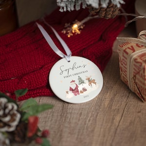 May include: A round white ceramic ornament with a white ribbon hanger. The front features the text "Sophia's FIRST CHRISTMAS" above a watercolor illustration of Santa Claus, a reindeer, a Christmas tree, gifts, and the year 2023.