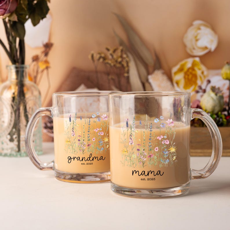 May include: Two clear glass mugs with handles, filled with a light brown liquid. Each mug features a floral design with the words "grandma est. 2025" and "mama est. 2025" in black script. The mugs are decorated with colorful wildflowers.