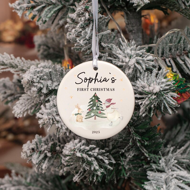 May include: A round Christmas ornament with the text "Sophia's First Christmas" and the year 2025. The ornament features a watercolor illustration of a snowman, a bunny, and a Christmas tree. The ornament hangs from a white ribbon.