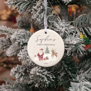 May include: A round white ceramic Christmas ornament hanging from a thin white ribbon. The ornament features the text Sophia's FIRST CHRISTMAS and 2025, with a printed illustration of Santa Claus, a small pine tree, a reindeer, and gift boxes.