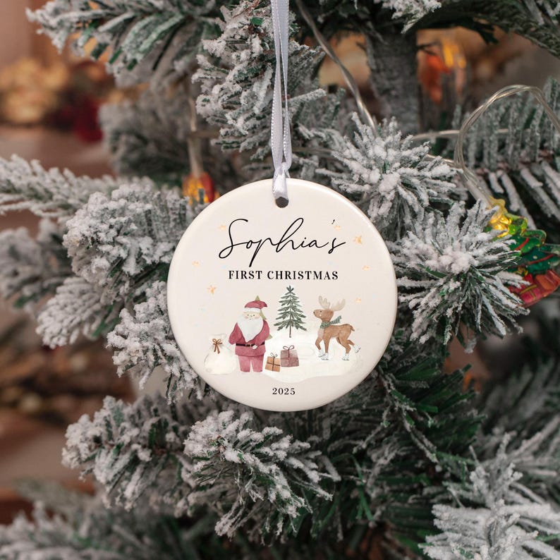 May include: A round white ceramic Christmas ornament hanging from a thin white ribbon. The ornament features the text Sophia's FIRST CHRISTMAS and 2025, with a printed illustration of Santa Claus, a small pine tree, a reindeer, and gift boxes.