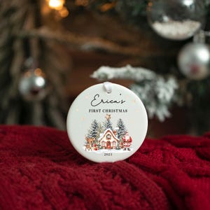 May include: A round white ceramic Christmas ornament featuring the text Erica's FIRST CHRISTMAS 2025. The design includes a small house, a reindeer, and Santa Claus surrounded by snow-covered pine trees and a star, hanging by a white ribbon.