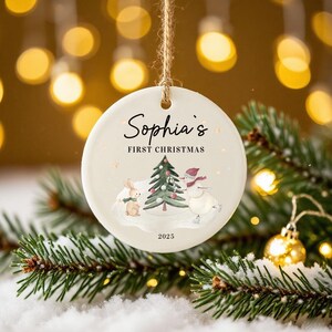 May include: A round, cream-colored Christmas ornament with the text "Sophia's FIRST CHRISTMAS" in black script. The ornament features a cartoon illustration of a bunny, a Christmas tree, and a snowman. The year "2023" is printed at the bottom. The ornament hangs from a twine string.