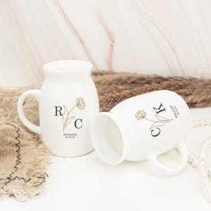 May include: Two white ceramic mugs with gold floral designs and personalized initials. The mug on the left has the initials "RC" and the text "ENGAGED 09.09.24". The mug on the right has the initials "CK" and the text "WEDDING ANNIVERSARY".