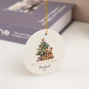 May include: A round white ceramic ornament hanging from a gold string. The front features a watercolor illustration of a teddy bear sitting next to a decorated Christmas tree with a star on top. Text reads My First Christmas, Michael, and 2025.