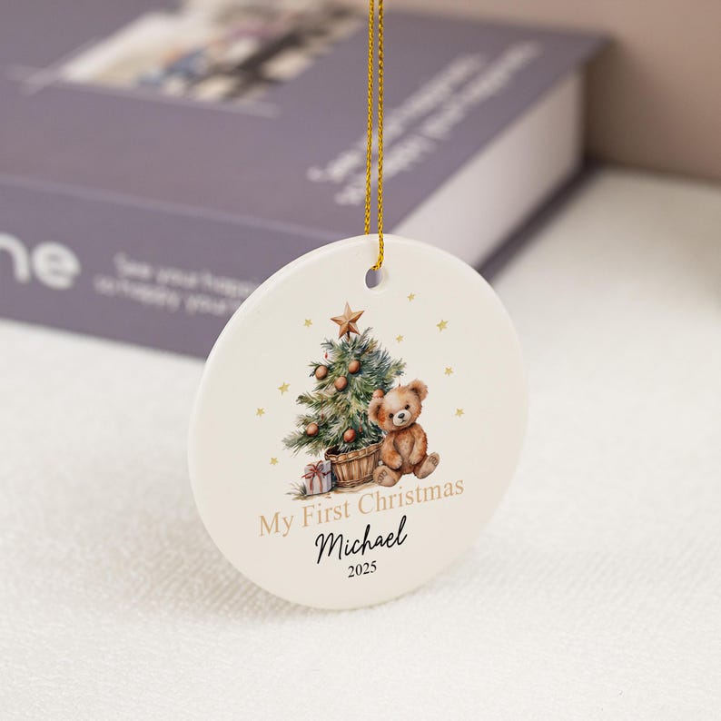 May include: A round white ceramic ornament hanging from a gold string. The front features a watercolor illustration of a teddy bear sitting next to a decorated Christmas tree with a star on top. Text reads My First Christmas, Michael, and 2025.