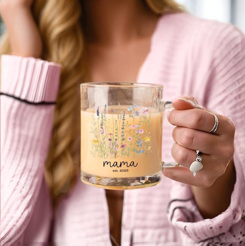 May include: A clear glass mug filled with a light-colored beverage. The mug features a floral design with the word "mama" and the text "est. 2025" in black. The mug has a handle and is held by a person.