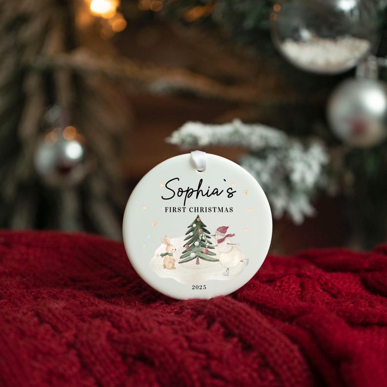 May include: A round Christmas ornament with the text "Sophia's First Christmas" and the year 2025. The ornament features a watercolor illustration of a snowman, a rabbit, and a Christmas tree. The ornament is white with a ribbon for hanging.