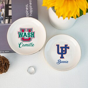 Puede incluir: Dos platos de cer&aacute;mica blancos para baratijas con bordes dorados. Un plato presenta un logotipo verde y rojo con la palabra "WASH" y el nombre "Camille". El otro plato tiene un logotipo azul y el nombre "Bonnie". Tambi&eacute;n se ven un anillo y un arreglo floral.