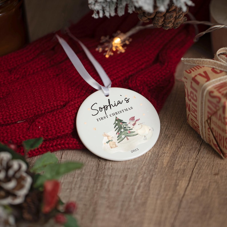 May include: A white ceramic Christmas ornament with the text "Sophia's First Christmas" and a watercolor illustration of a Christmas tree, snowman, and bunny. The ornament is hanging on a red knitted surface.
