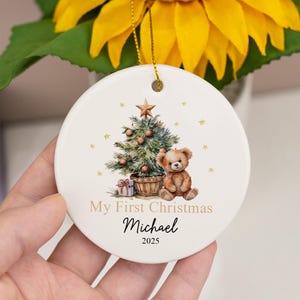 May include: A round white ceramic ornament featuring a printed illustration of a decorated Christmas tree, a brown teddy bear, and a gift box. The text reads My First Christmas, the name Michael, and the year 2025 in black and gold lettering.
