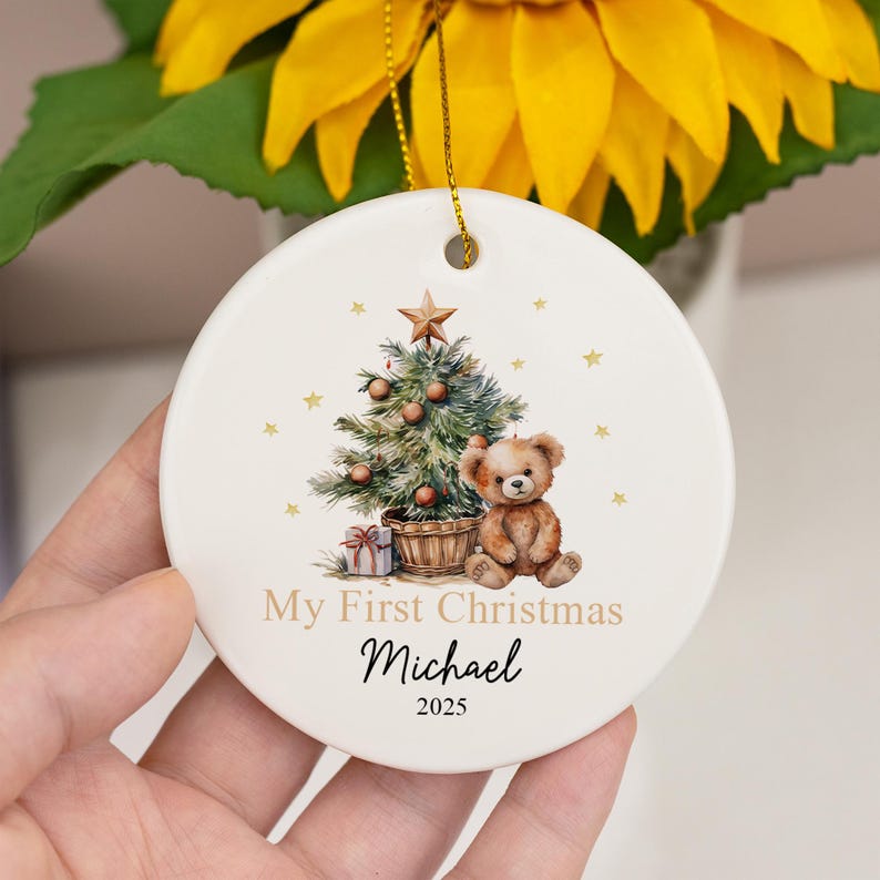 May include: A round white ceramic ornament featuring a printed illustration of a decorated Christmas tree, a brown teddy bear, and a gift box. The text reads My First Christmas, the name Michael, and the year 2025 in black and gold lettering.