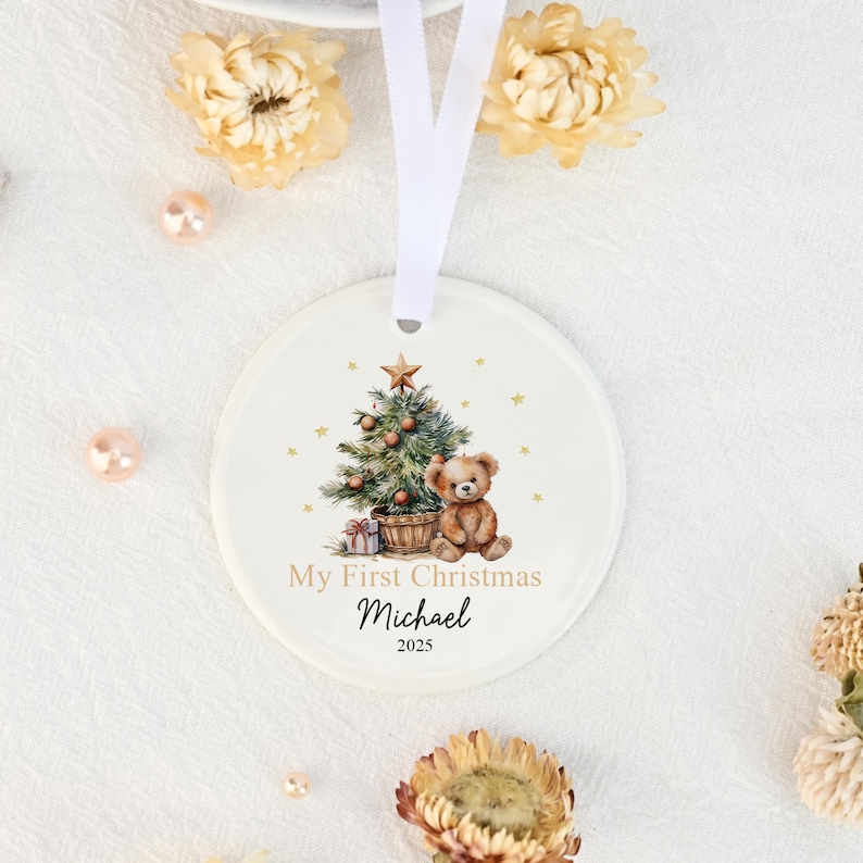May include: A round white ceramic ornament featuring a watercolor illustration of a teddy bear sitting next to a decorated Christmas tree. The text reads My First Christmas, the name Michael, and the year 2025. It hangs from a white ribbon.