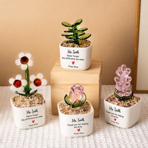 Puede incluir: Cuatro pequeñas macetas de cerámica blanca con plantas decorativas de vidrio. Una tiene flores tipo margarita, otra una suculenta y dos plantas tipo cristal. Cada maceta tiene un mensaje y un símbolo de corazón.