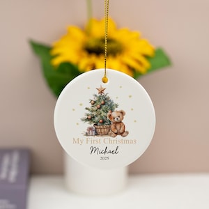 May include: A round white ceramic ornament hanging from a gold string. The design features a decorated Christmas tree, a brown teddy bear, and small gold stars. The text reads My First Christmas, the name Michael, and the year 2025.