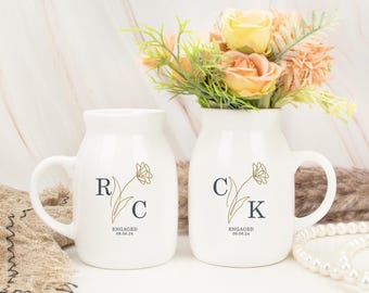 May include: Two white ceramic mugs featuring a gold line-art flower graphic. The left mug displays the letters R and C, while the right mug displays C and K. Both mugs include the text ENGAGED 09.08.24 printed in a dark gray serif font.