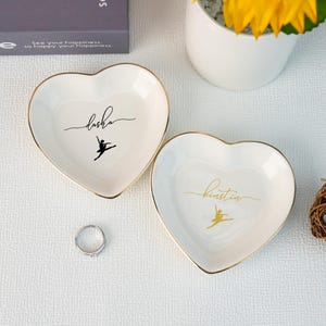 Könnte beinhalten: Zwei weiße herzförmige Keramik-Schmuckschalen mit goldener Einfassung. Eine Schale trägt den Namen "Dasha" in schwarzer Schrift mit einer schwarzen Silhouette einer Ballerina. Die andere Schale trägt den Namen "Kristin" in goldener Schrift mit einer goldenen Silhouette einer Ballerina.