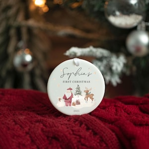 May include: A round white ceramic Christmas ornament featuring the text Sophia's FIRST CHRISTMAS 2025. The design includes a watercolor illustration of Santa Claus, a small Christmas tree, a reindeer, and gift boxes on a snowy ground.