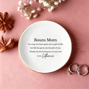以下が含まれることがあります： 金色の縁取りが施された白いセラミックプレート。テキスト「Bouns Mom」と心温まるメッセージが書かれています。 プレートは装飾的な花と2つの指輪に囲まれ、ピンク色の背景に配置されています。